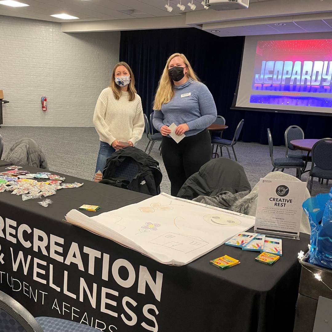 Image of two RecWell staff behind a Recreation & Wellness table in front of a Jeopardy screen.